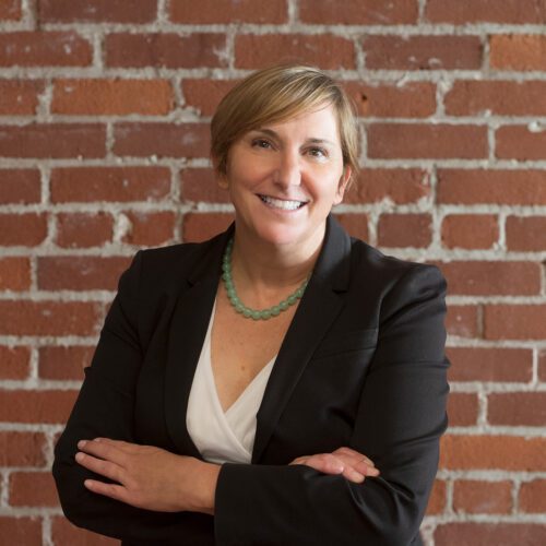 Darby Kernan wearing black jacket and white shirt and jade stone neckless standing in front of brick wall