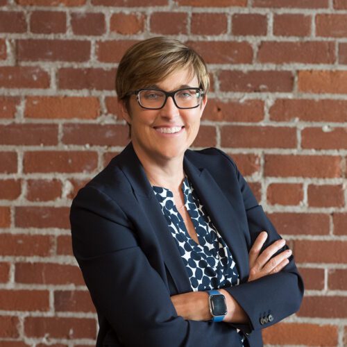 Kathy Mossburg wearing dark blue jacket and blue and whit shirt standing in front of brick wall