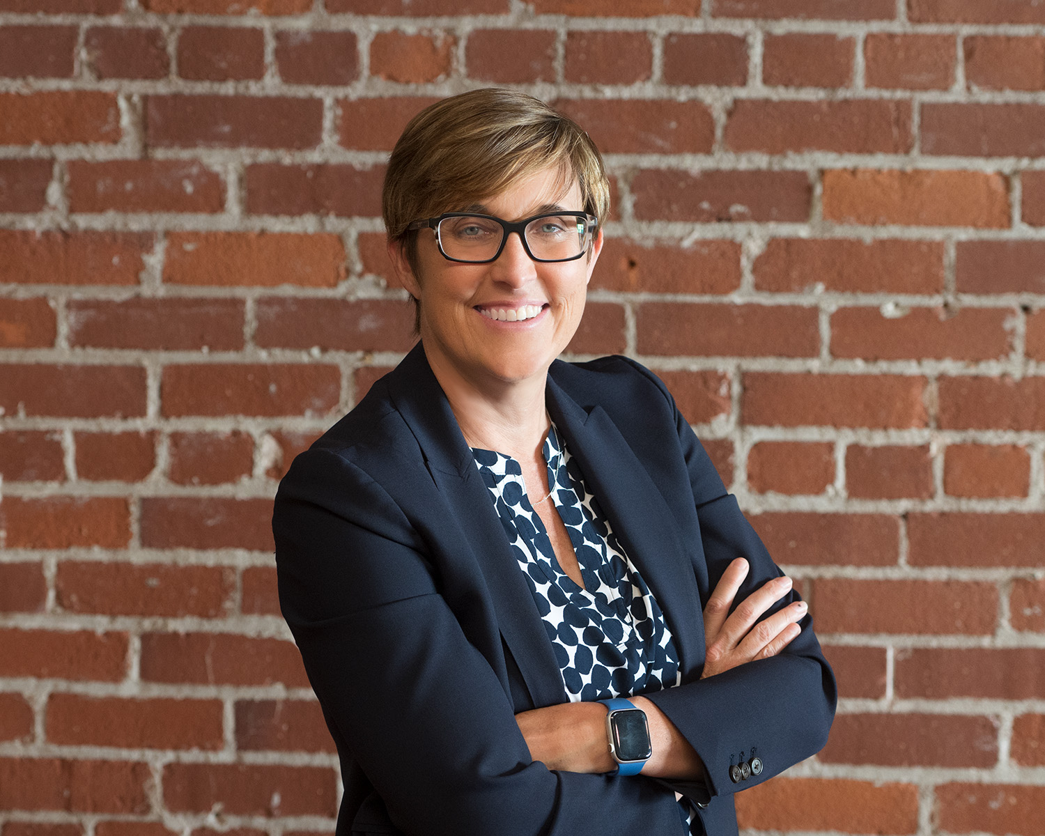 Kathy Mossburg wearing dark blue jacket and blue and whit shirt standing in front of brick wall
